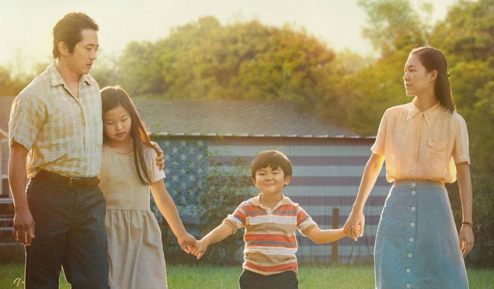 Still from "Minari" featuring a Korean family holding hands on a farm with an American flag in the background