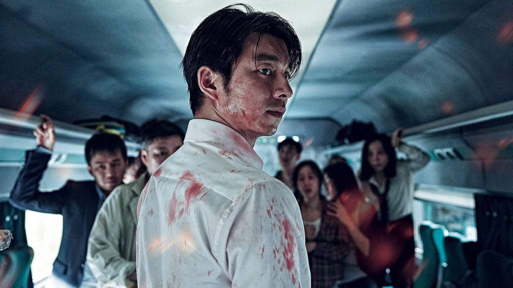 Still from "Train to Busan" featuring the main character covered in blood, looking over his shoulder in a train car with frightened people behind him
