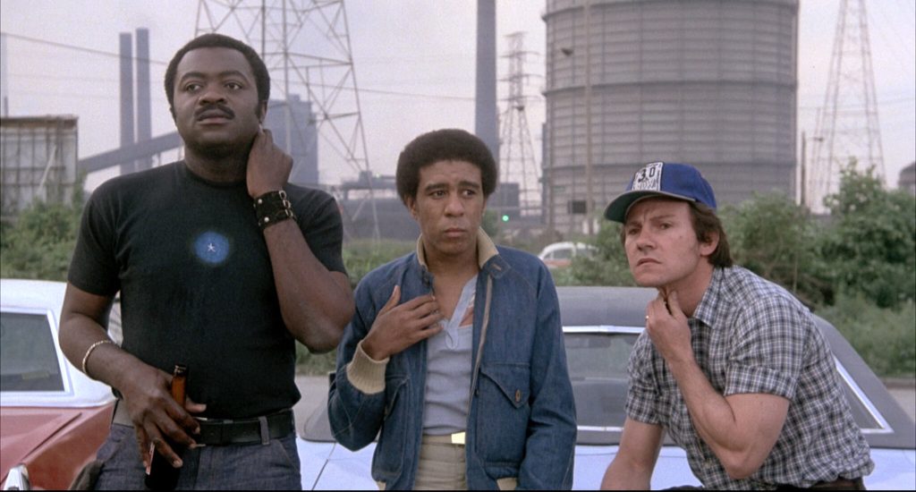 Still from "Blue Collar" featuring three men leaning on the hood of a car