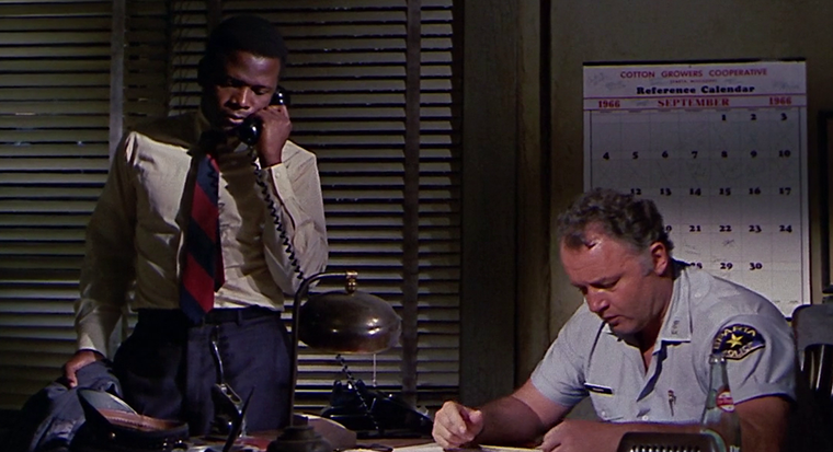 Still from "In the Heat of the Night" featuring a man leaning on a police officer's desk while talking on the phone
