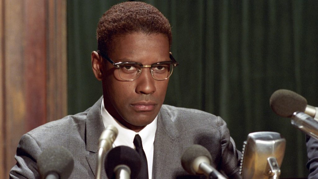 Still from "Malcom X" featuring Malcolm sitting in front of an array of microphones