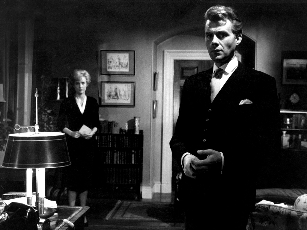 Still from "Victim" featuring a woman in the background gazing at a man in the foreground who is staring into the camera