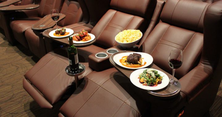 Brown, reclined movie theater seats are set with luxurious plates of scrumptious food and drink