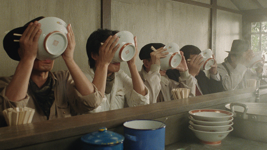 A row of five men dressed similarly in casual clothes sit at a food counter inside a restaurant. Each man is holding a bowl with chopsticks up to their mouths, seemingly drinking from their bowls. The bowls obstruct their faces