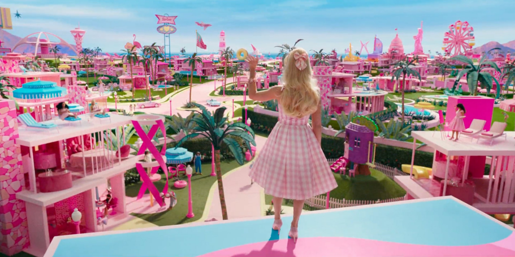 Main character Barbie is shown from behind and stands atop a dreamhouse wearing a pink checkered dress and matching pink bow in her hair. She looks out at a fictional, vibrant Barbieland decorated with pink houses and palmtrees and waves to her neighbors. 
