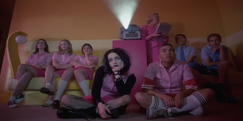 An orange room is darkened aside from the light of a projector glowing in the top center of the image. On either side of the projector sits teenage girls dressed in all pink and teenage boys dressed in all blue, seemingly watching whatever is projected in front of them. They all appear disinterested. 