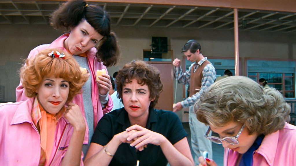 Three women sit together outside in a plaza, two wearing pink leather jackets and one wearing a black shit. A fourth woman stands behind them also in a pink leather jacket. Three of the four women are admiring something in the distance. A man in the background plays with a Yo-Yo.