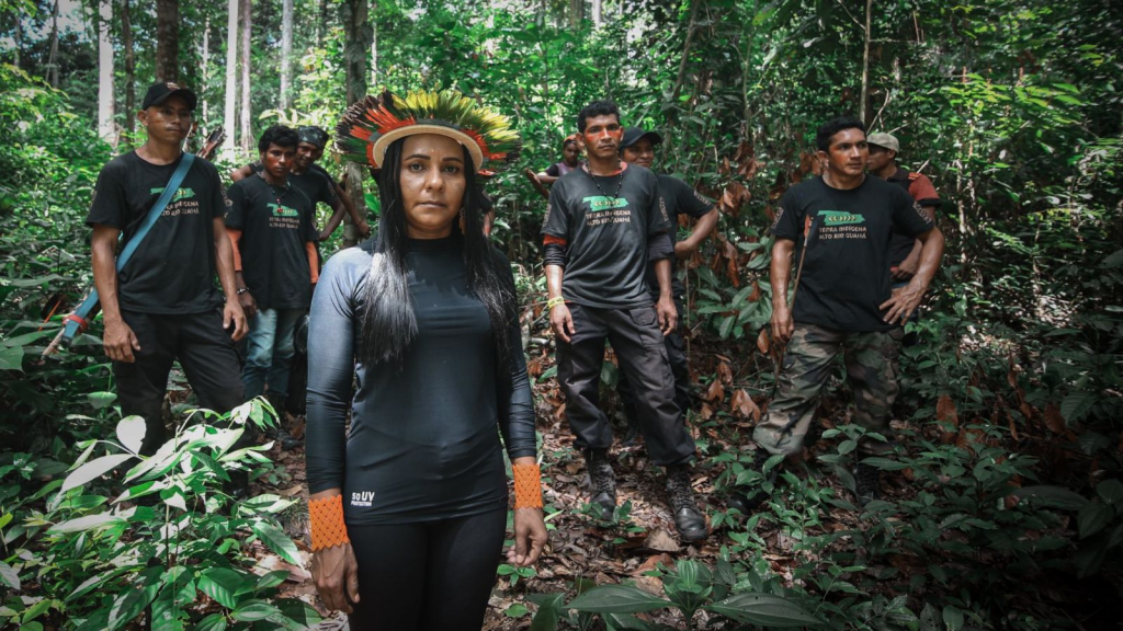 This is a photograph of a group of people standing in the Amazon Rainforest. They are all wearing black clothing and at the forefront of the group is a young woman wearing a halo-like headdress with multicolor feathers. Other members of the group are holding tools or have tools slung over their shoulder. A sunny sky lights up the leaves on trees around them.