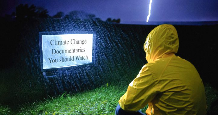 Person in a yellow rain jacket sits outside in a lightning storm looking at a TV that says "climate change documentaries you should watch."