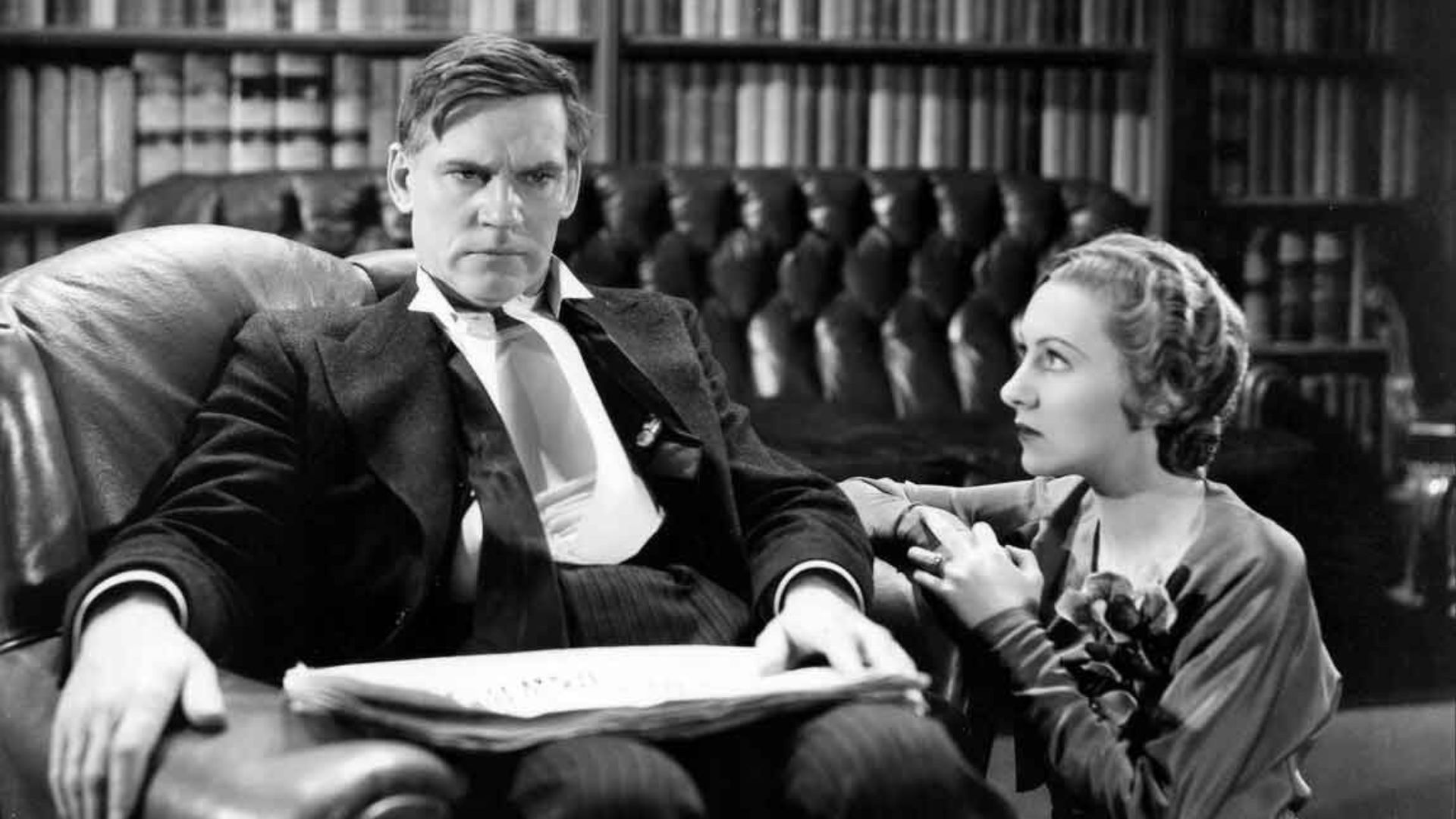A man in a suit sits in a chair with a woman sitting on the floor and looking at him.