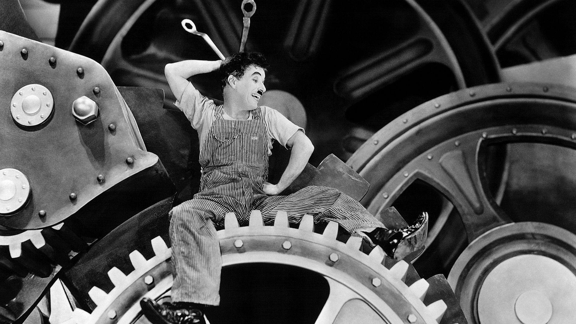 A man in pinstripe overalls sits on an oversized large metal gear.