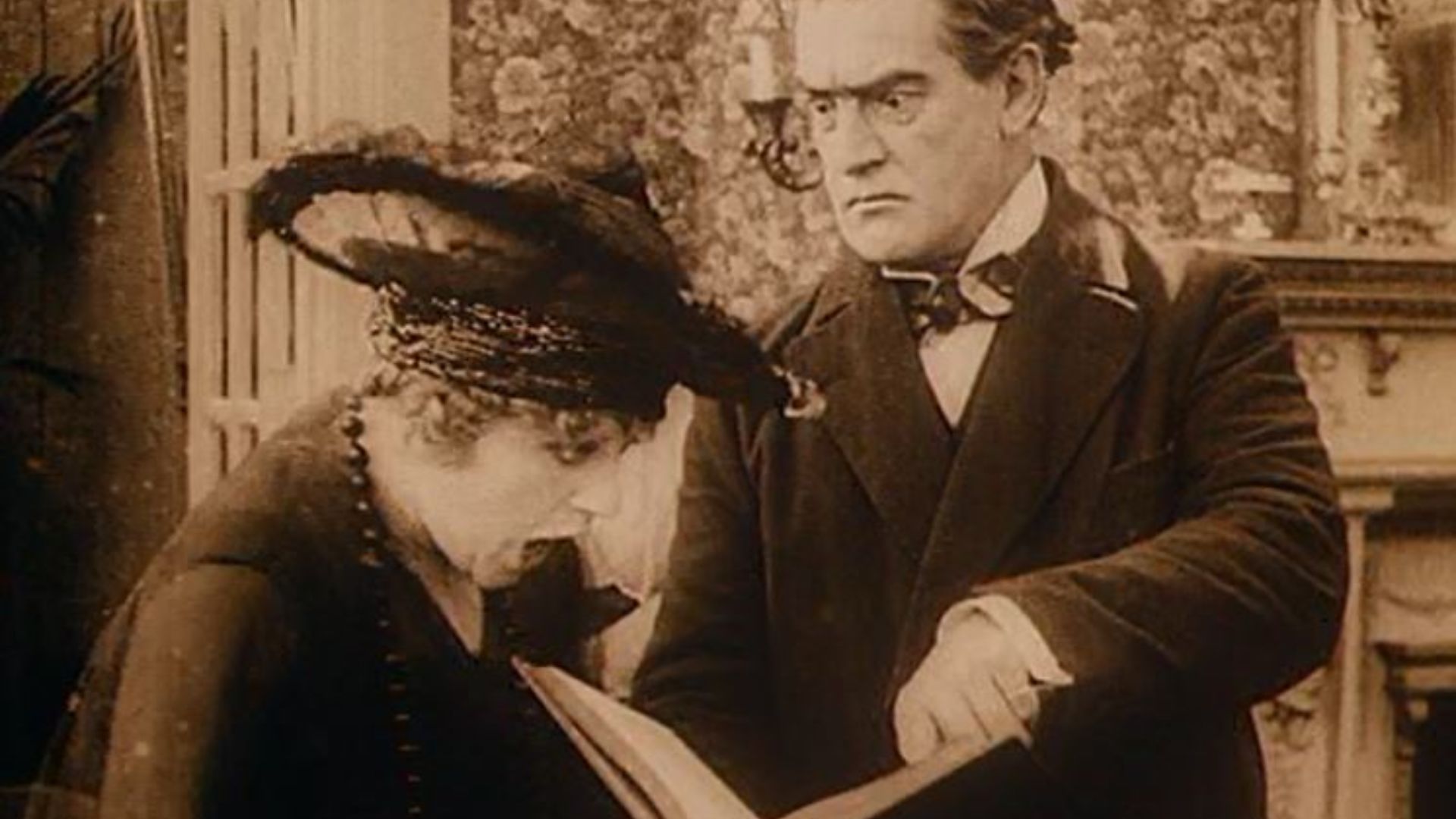 A woman in a large hat looks at a book, while a man in a suit stands with a concerned expression.