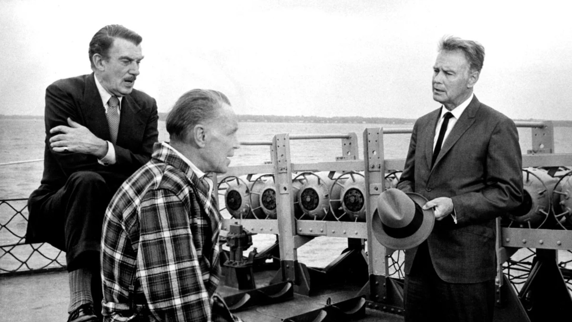 Three men, two seated, on the deck of a boat, with one carrying a hat in his hands.