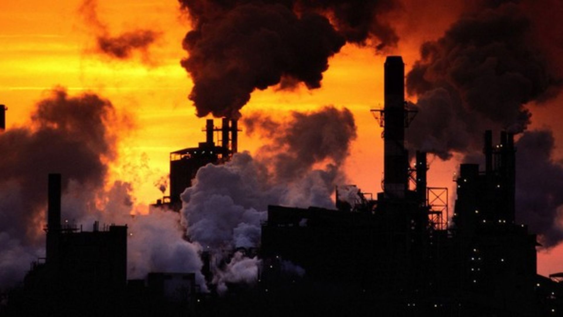 Silhouetted factories emit smoke against a bright orange sky.