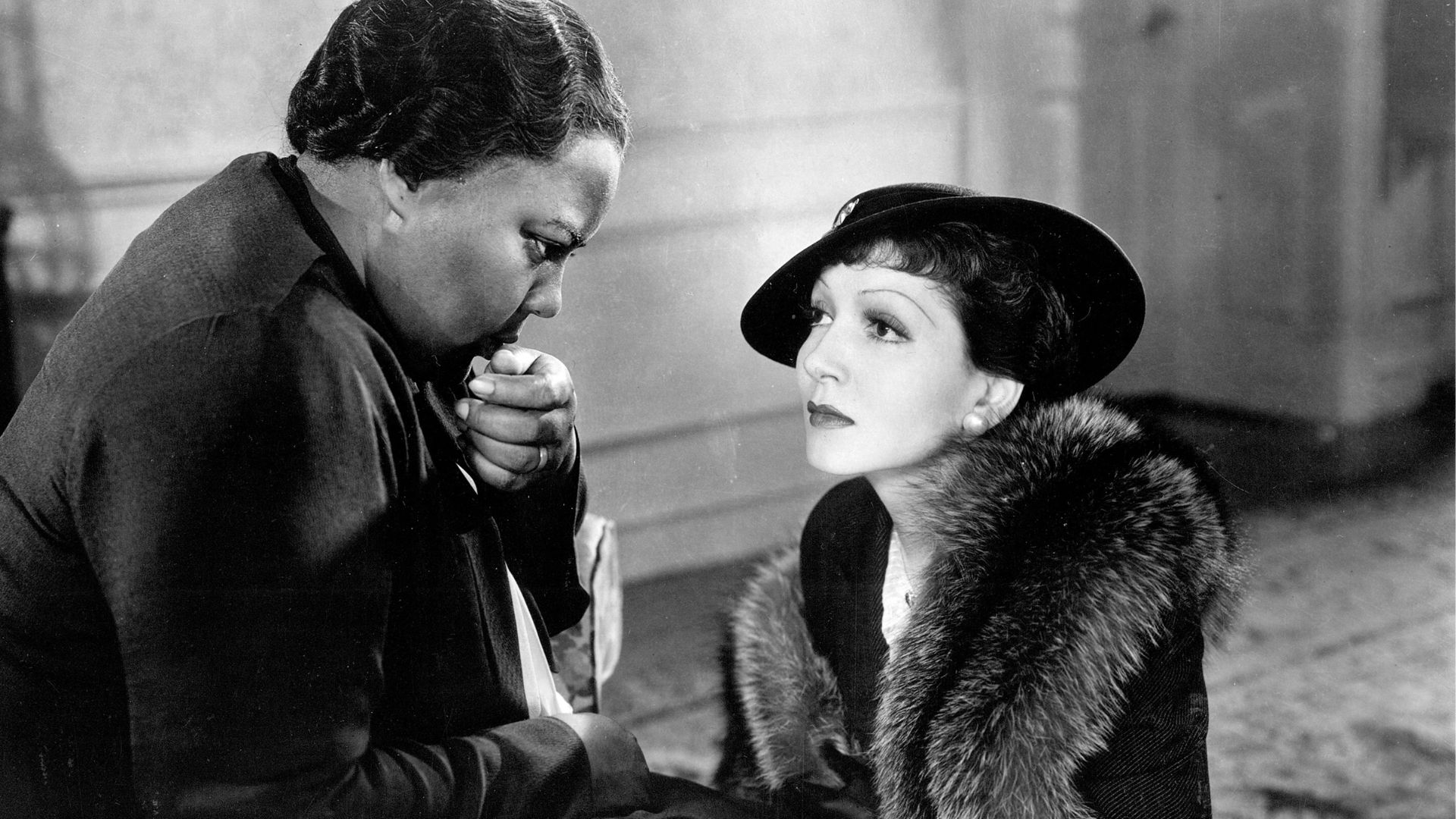 One woman sits in a chair, kissing the hand of a woman sitting on the floor inside a house.