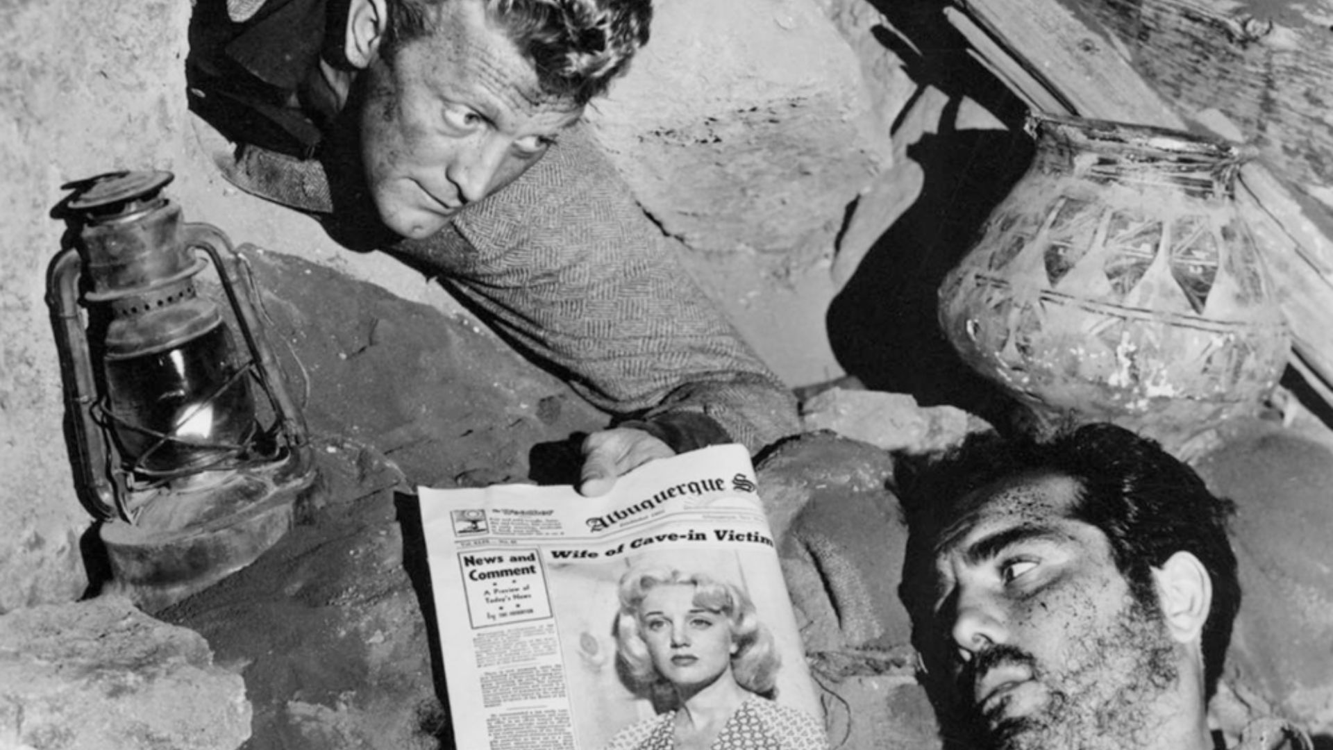 A scene of a man crawling down, and gazing down at another man lying on the floor next to a newspaper.