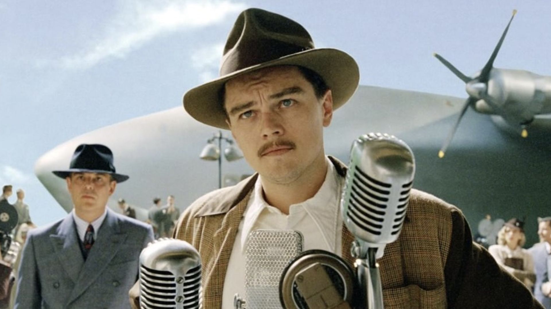 A man in a hat speaks into microphones against the background of a large blimp.