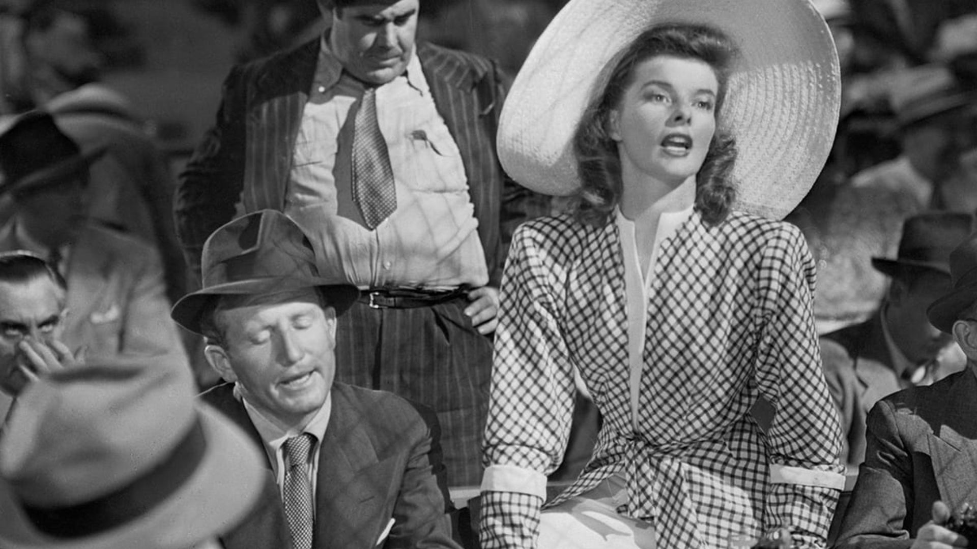 A woman wearing a large hat and patterned dress sits in front of a crowd of men.