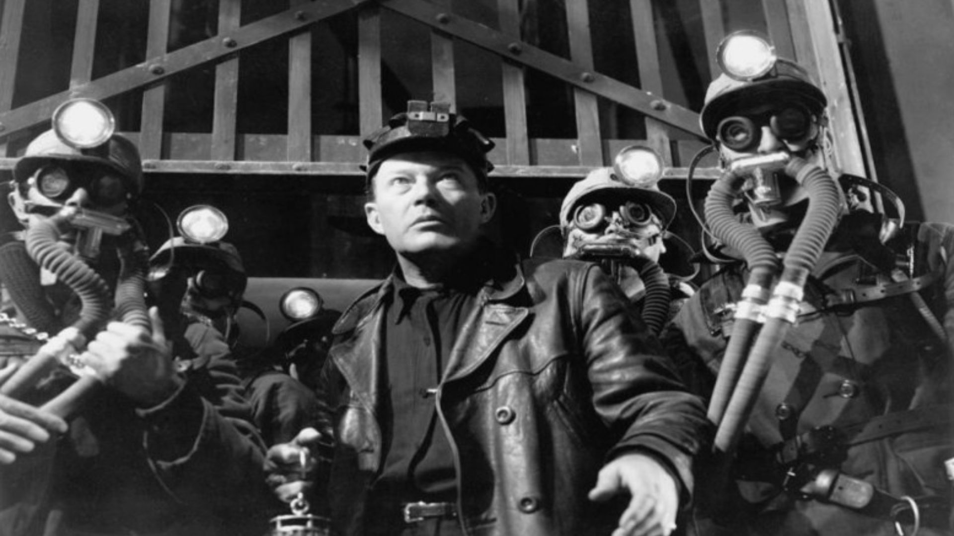A low shot of a group of coal miners wearing breathing masks standing in front of a large metal gate. One man stands in the middle without a mask.