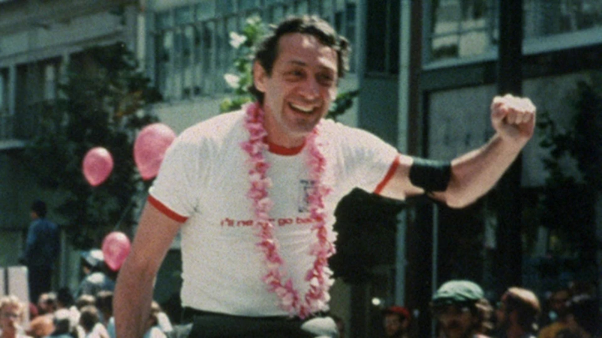 Harvey Milk, wearing a lei, smiles and holds up a fist against a parade background.