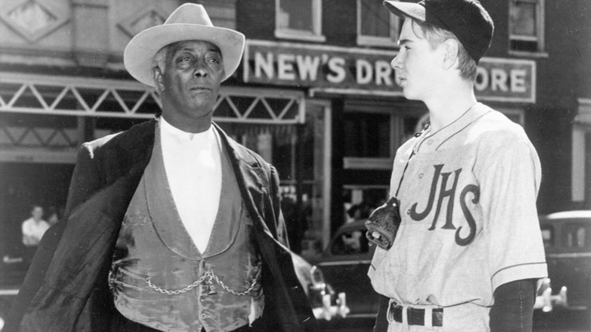 A black man in formal attire talks to a younger white man in a baseball uniform.