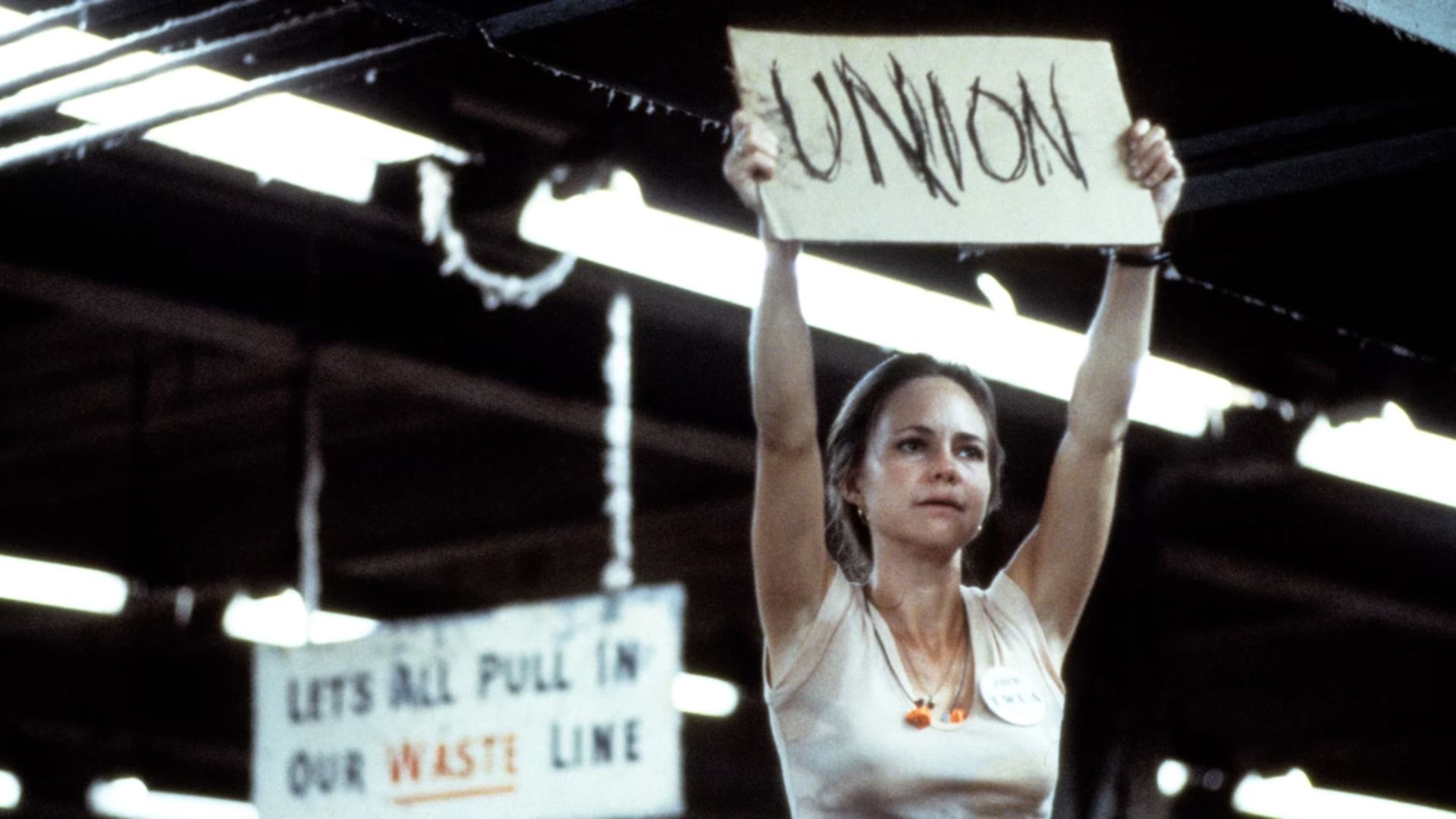 A woman holds up a sign reading "UNION", in front of a sign hanging from the ceiling that reads "LET'S ALL PULL IN OUR WASTE LINE".