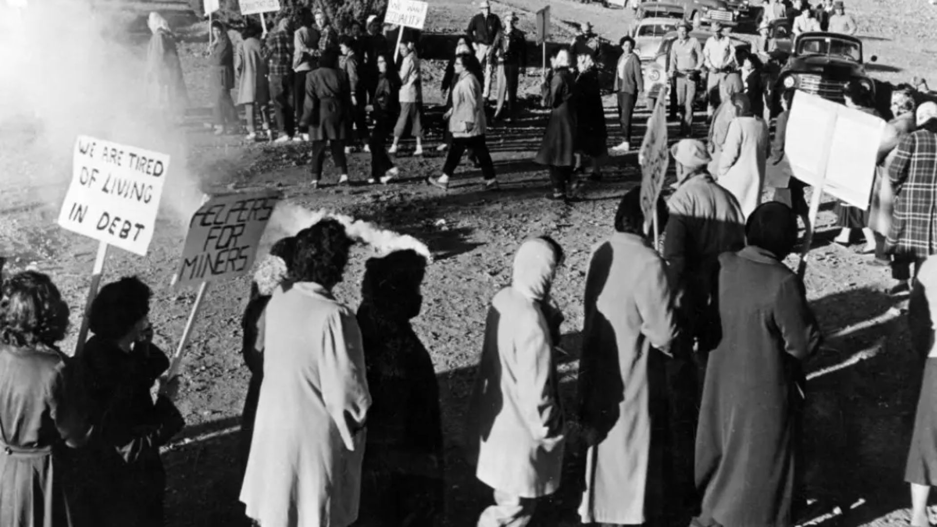 A group of women, protests with signs that read "WE ARE TIRED OF LIVING IN DEBT" and "CLEAN UP FOR MINERS."