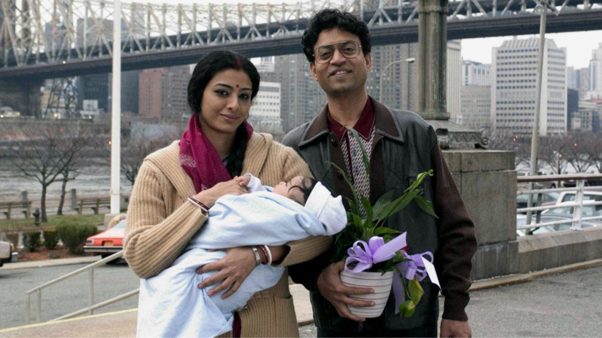 A young woman, holding an infant, and a young man, holding a small plant, stand side by side in front of a large bridge in New York City.