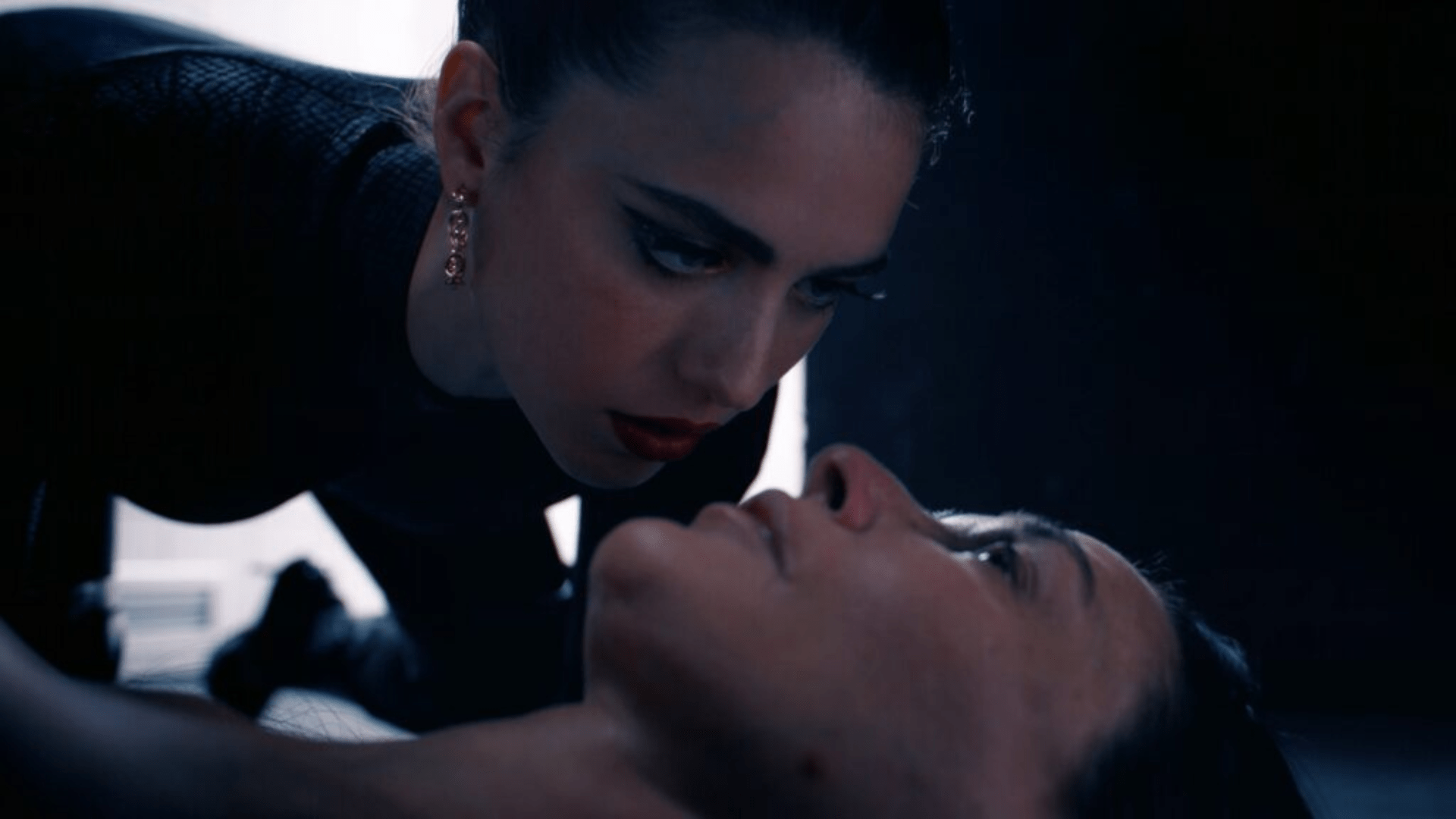 A younger woman hovers over the face of an older woman lying on the floor of a dark room.