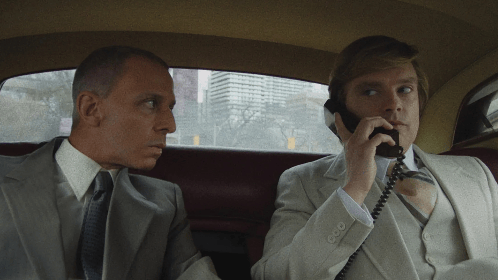 Two men sit in the backseat of a car. The older man on the left, gazes intently at the younger man, who is in the middle of a telephone call.