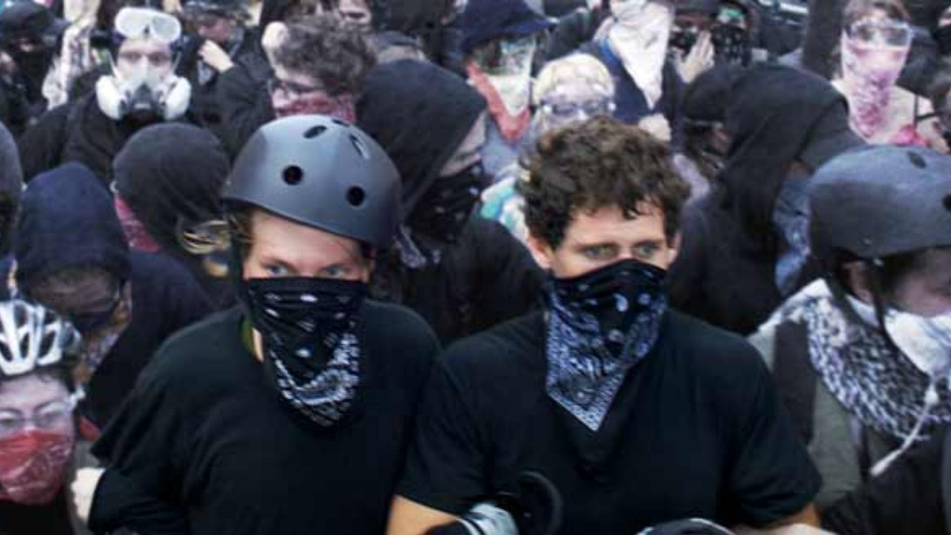 Two men with bandannas covering their mouths stand at the front of a crowd dressed in black.