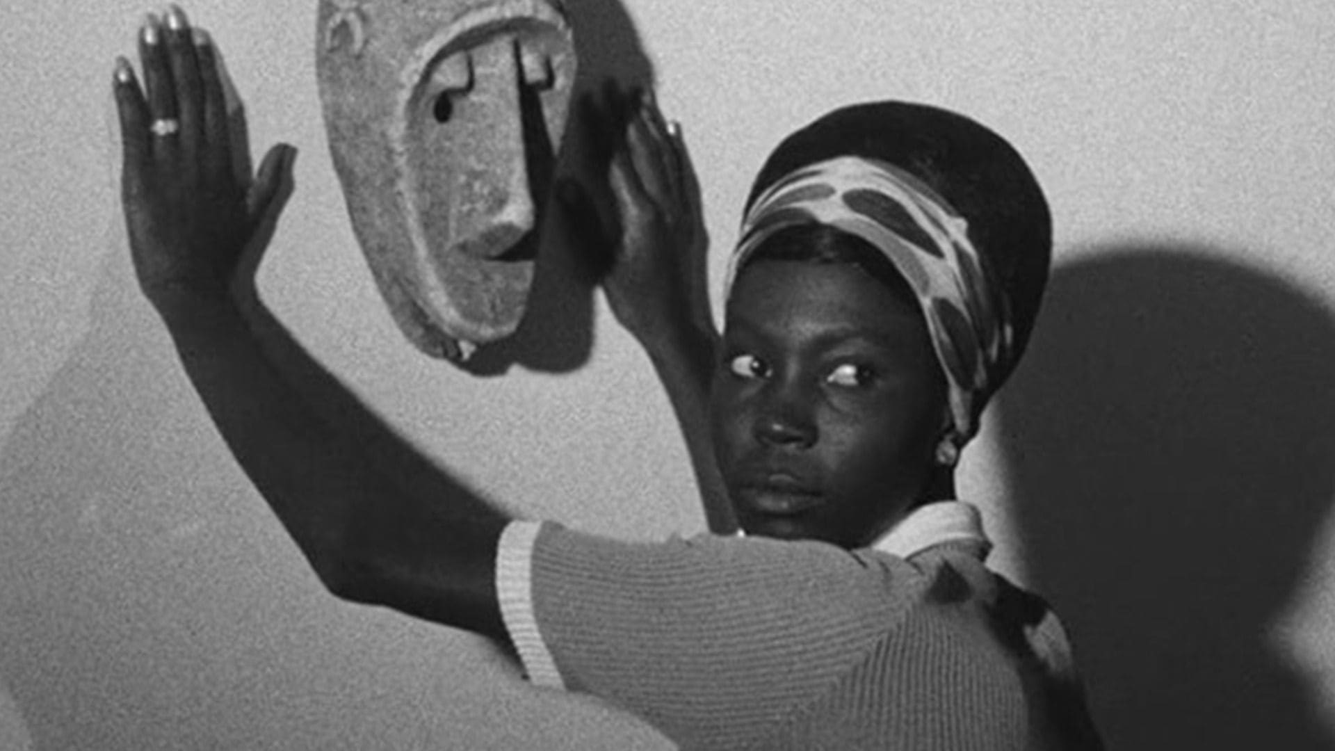 A woman looks behind her and stands with her hands flat on a wall, which a mask hangs on.