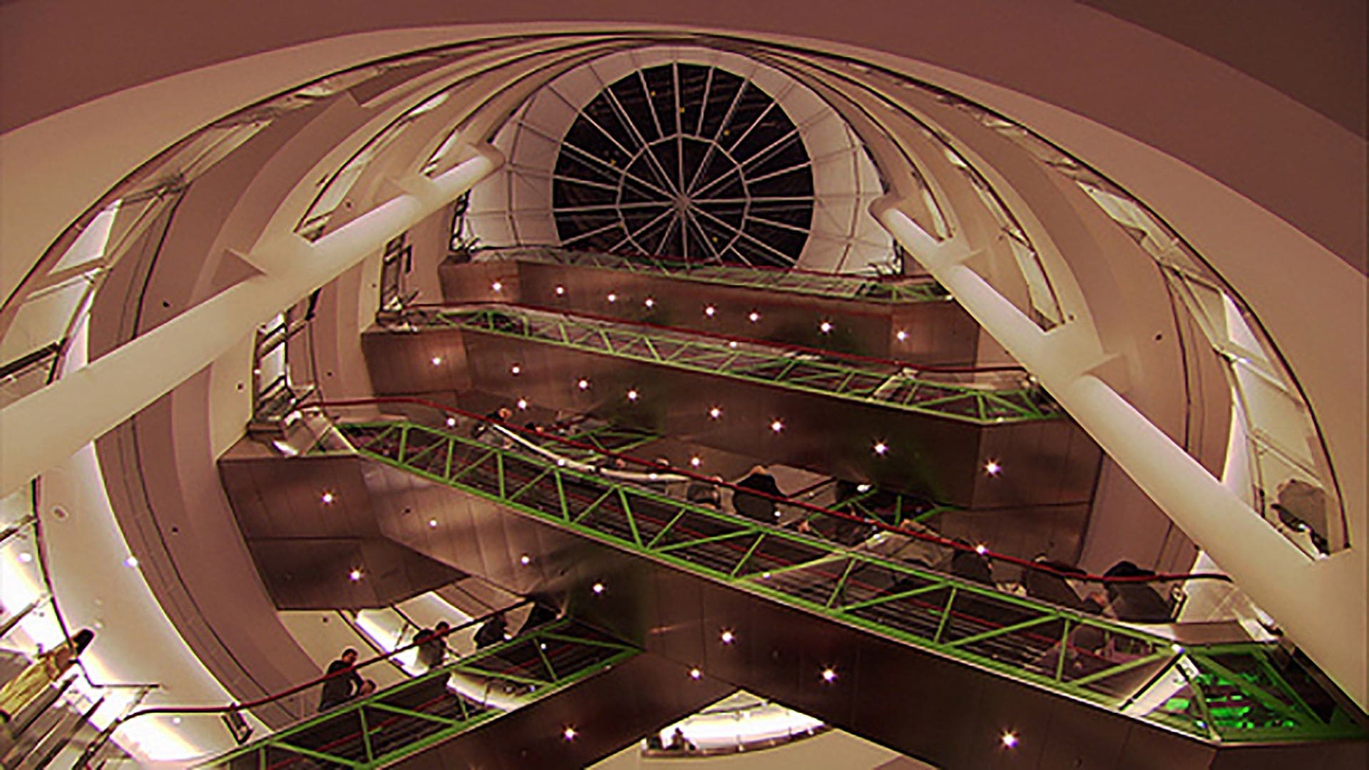 A view of a mall from the bottom floor, with green escalators and a skylight.