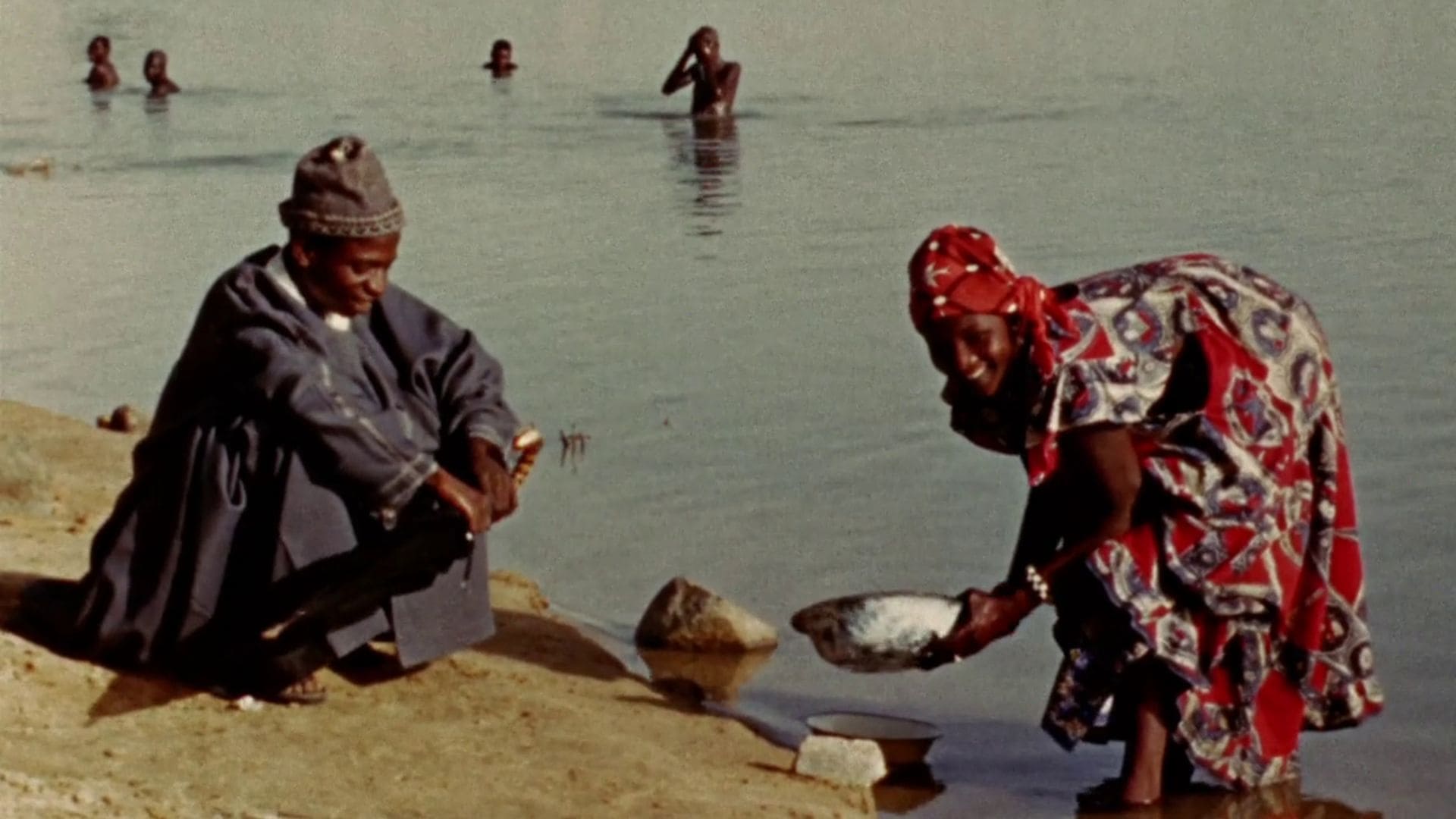 A man sits by the shore of a body of water while a woman next to him holds a bowl, crouched in the water.