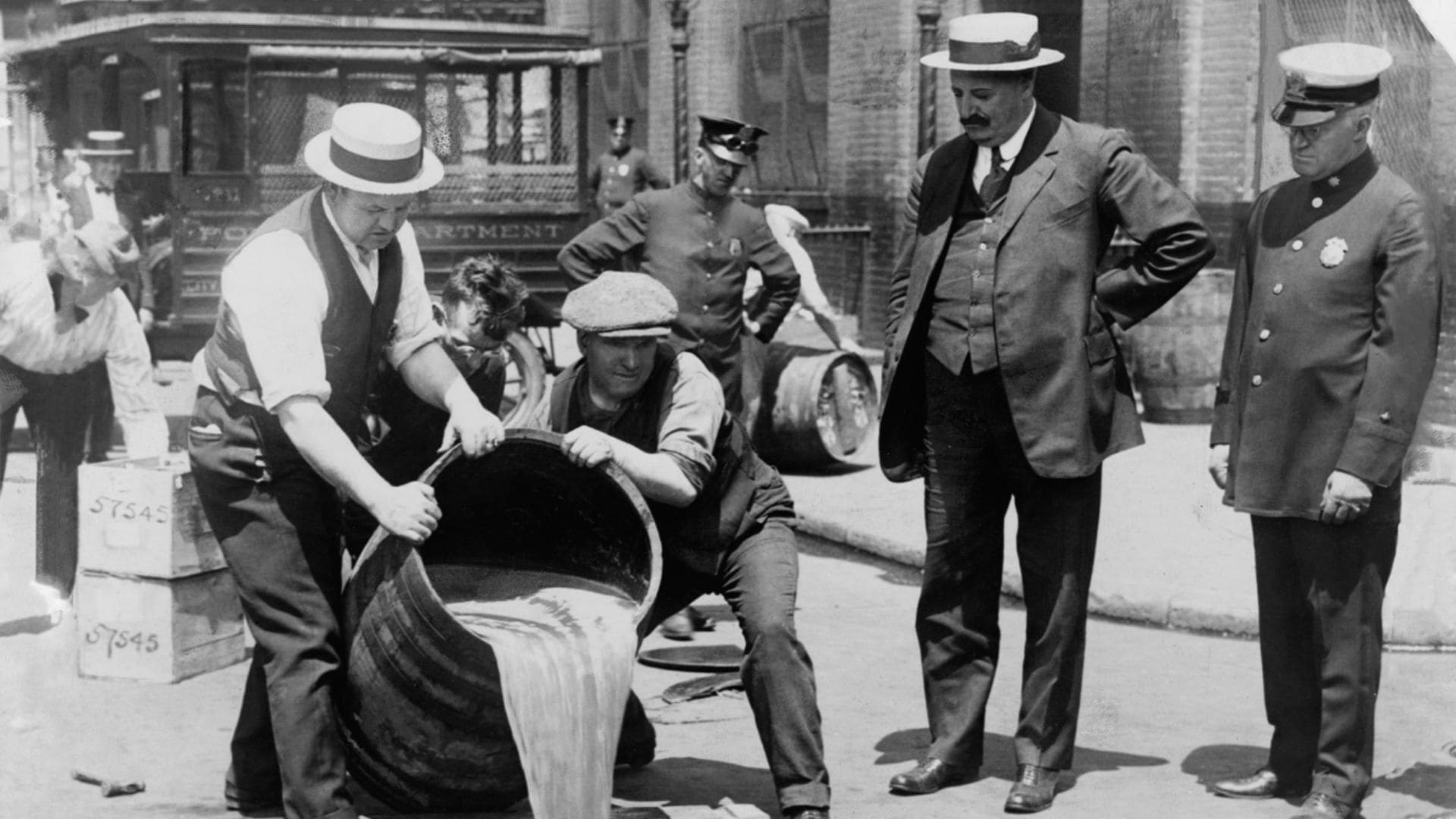 Two men pour a large barrel of liquid onto the street while two police officers and a man in a suit watch.