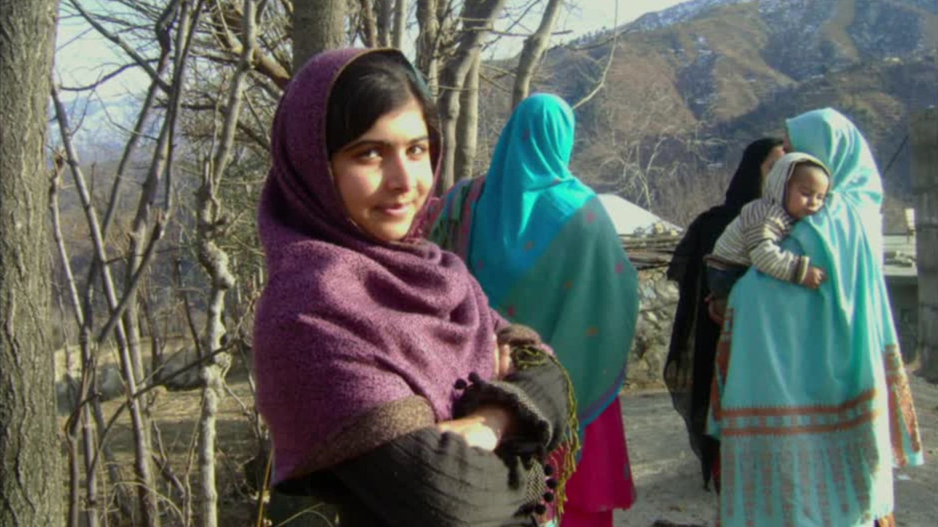 Activist Malala Yousafzai stands with a proud expression and her arms crossed in front of a crowd of women and children.
