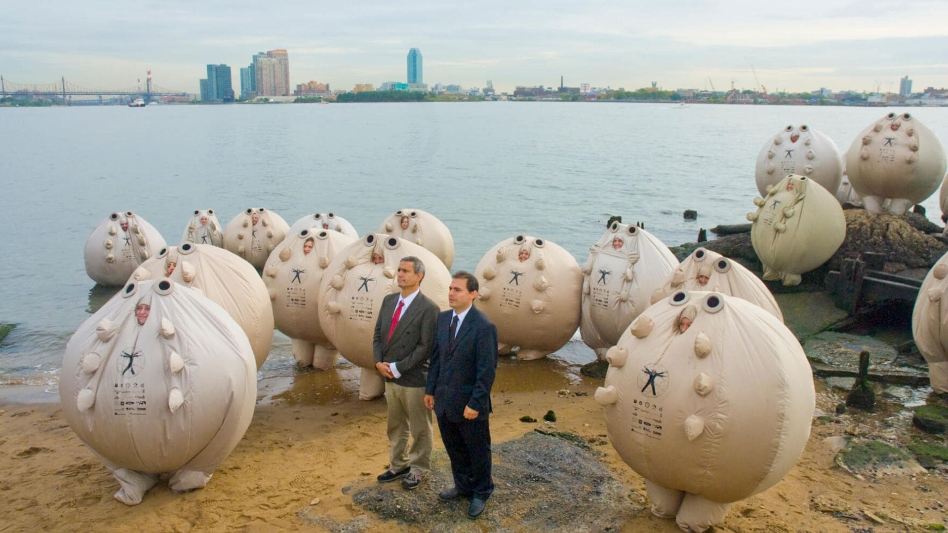 Two men stand amongst figures in inflatable costumes, on the shore of a body of water.