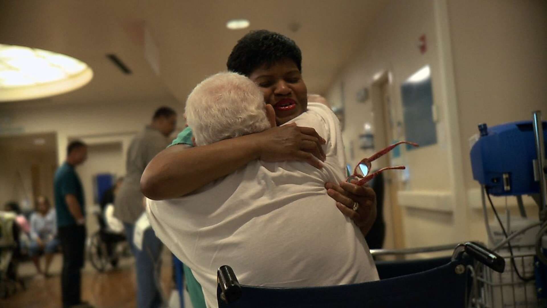 A nurse hugs a woman in a wheelchair in a hospital setting.