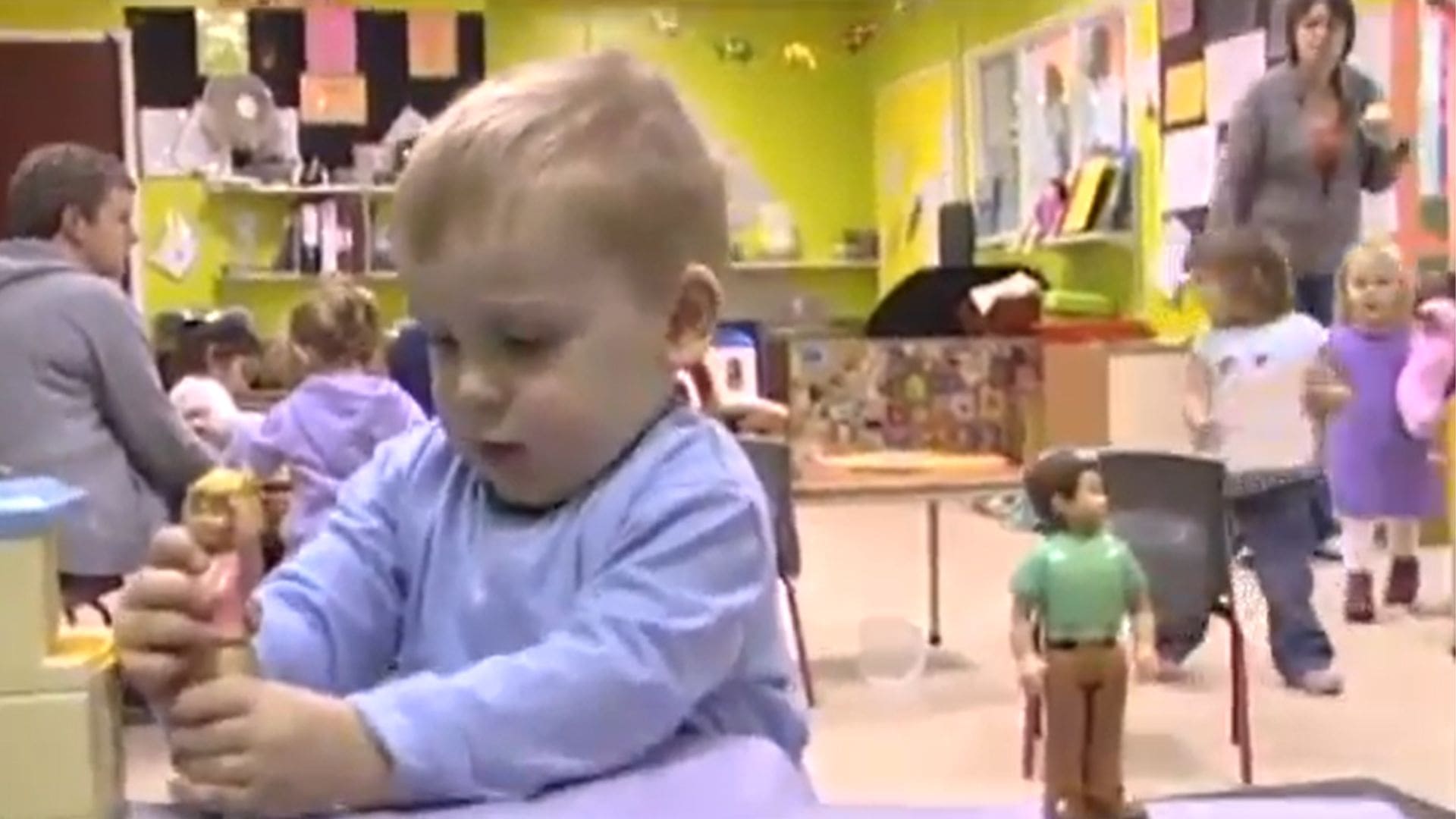 A baby plays with dolls in a room filled with other babies and toddlers doing the same.