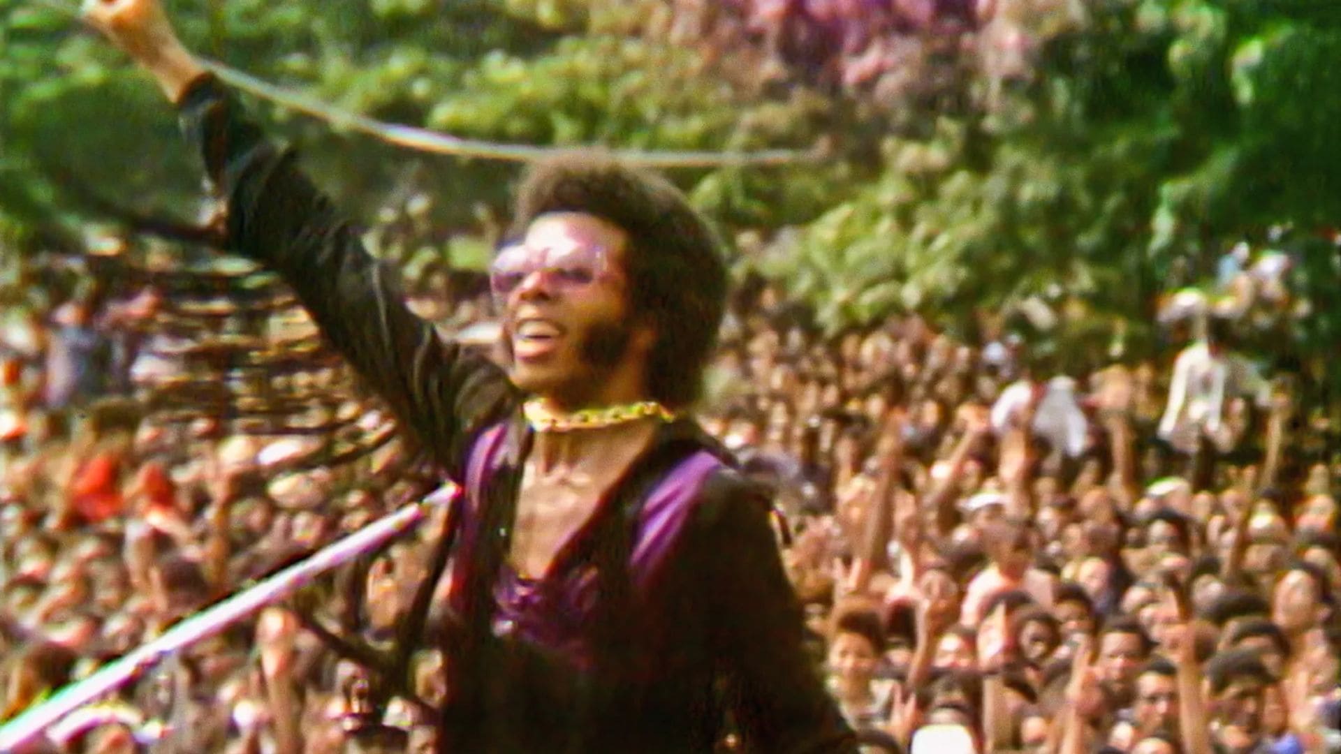Musician Sly Stone stands in front of a crowd with his fist in the air.