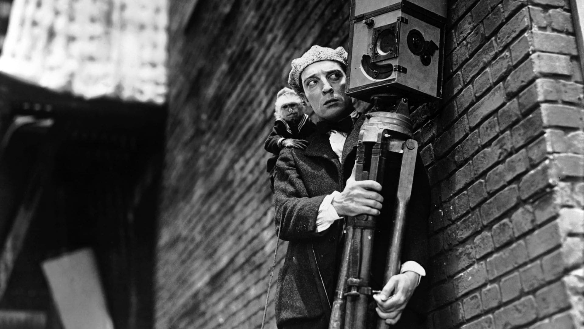 A man stands against a wall holding a camera taller than himself, and a small monkey perched on his back.