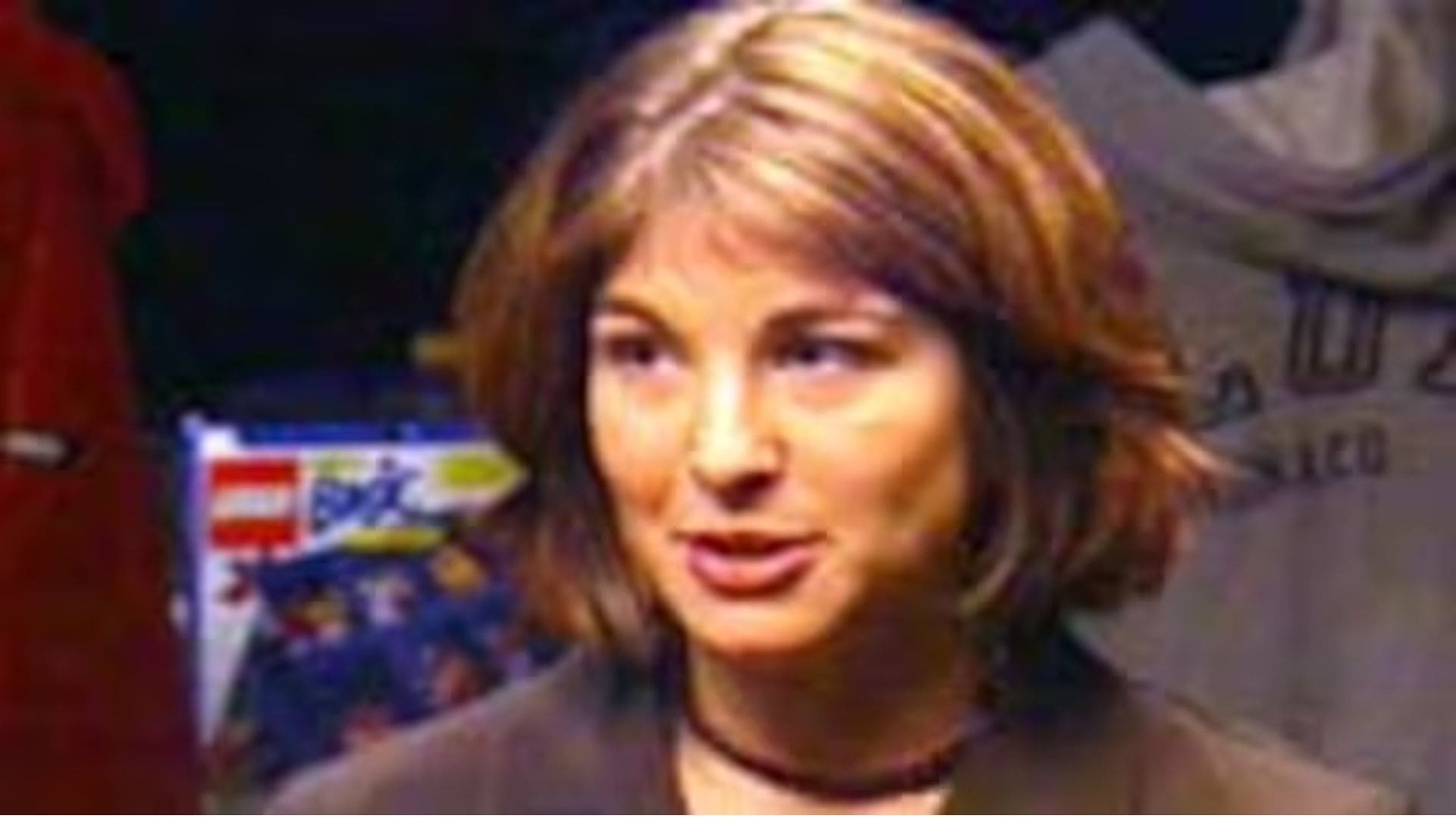 A grainy image of a woman speaking, with a packaged Lego set behind her.