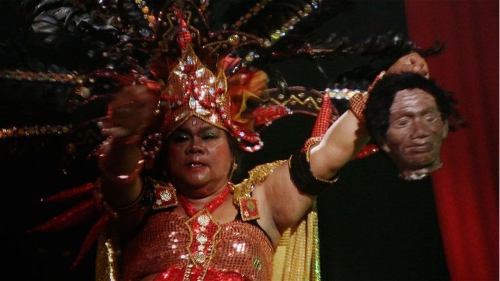 A woman in an elaborate headdress holds a prop, designed to look like a man's head.