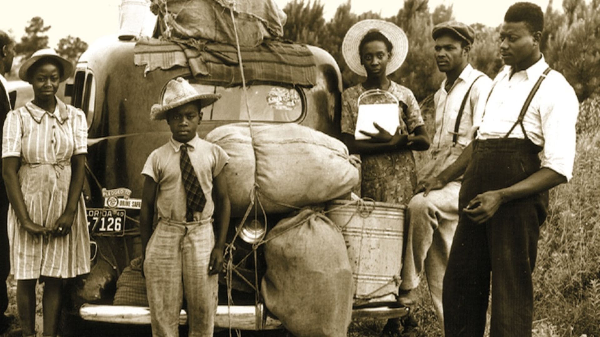 A black-and-white photo of a family standing around their car, which is packed with bags, buckets, and blankets.