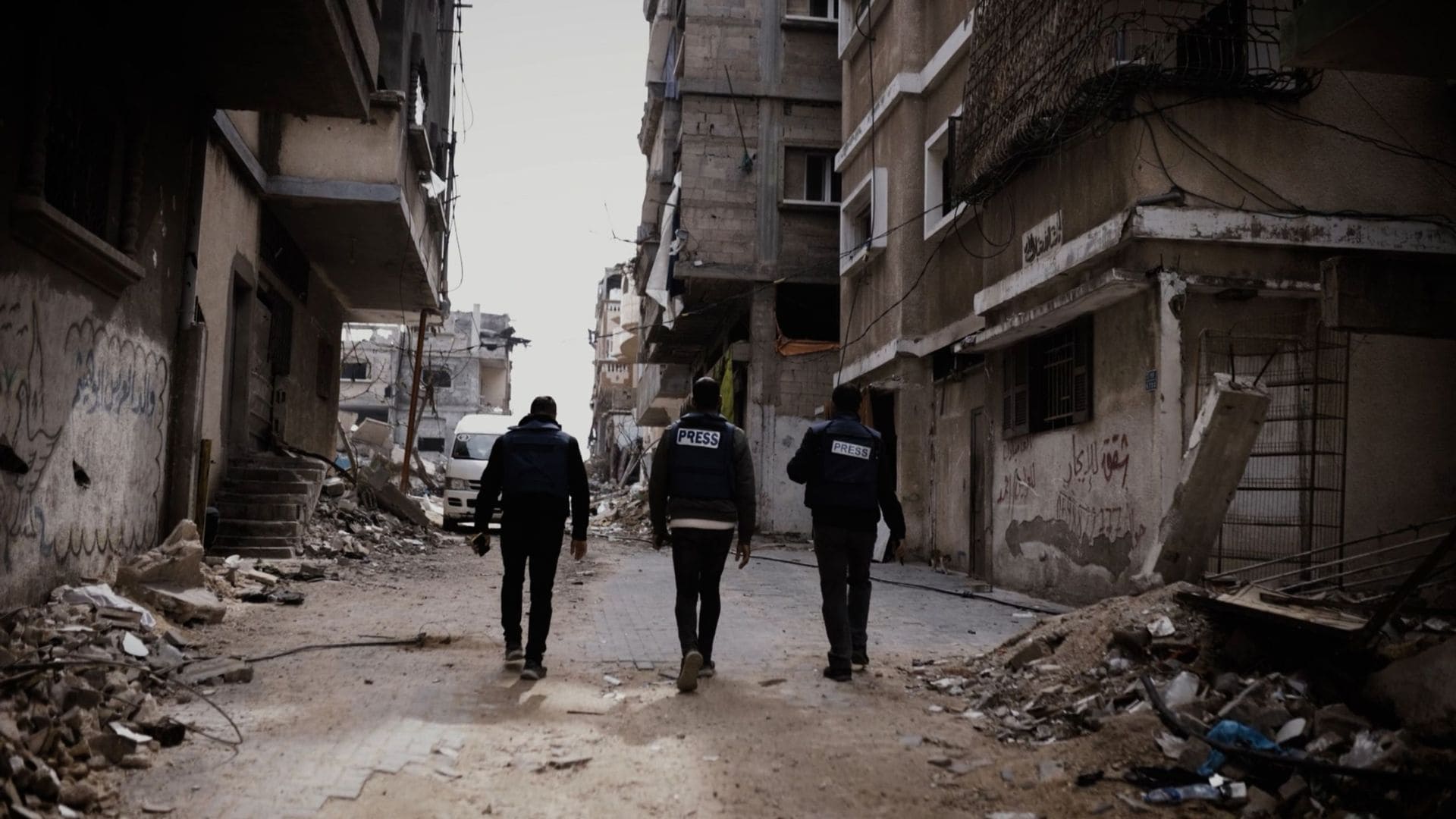 Three men walk the city of Gaza, with ruined buildings visible in the distance.