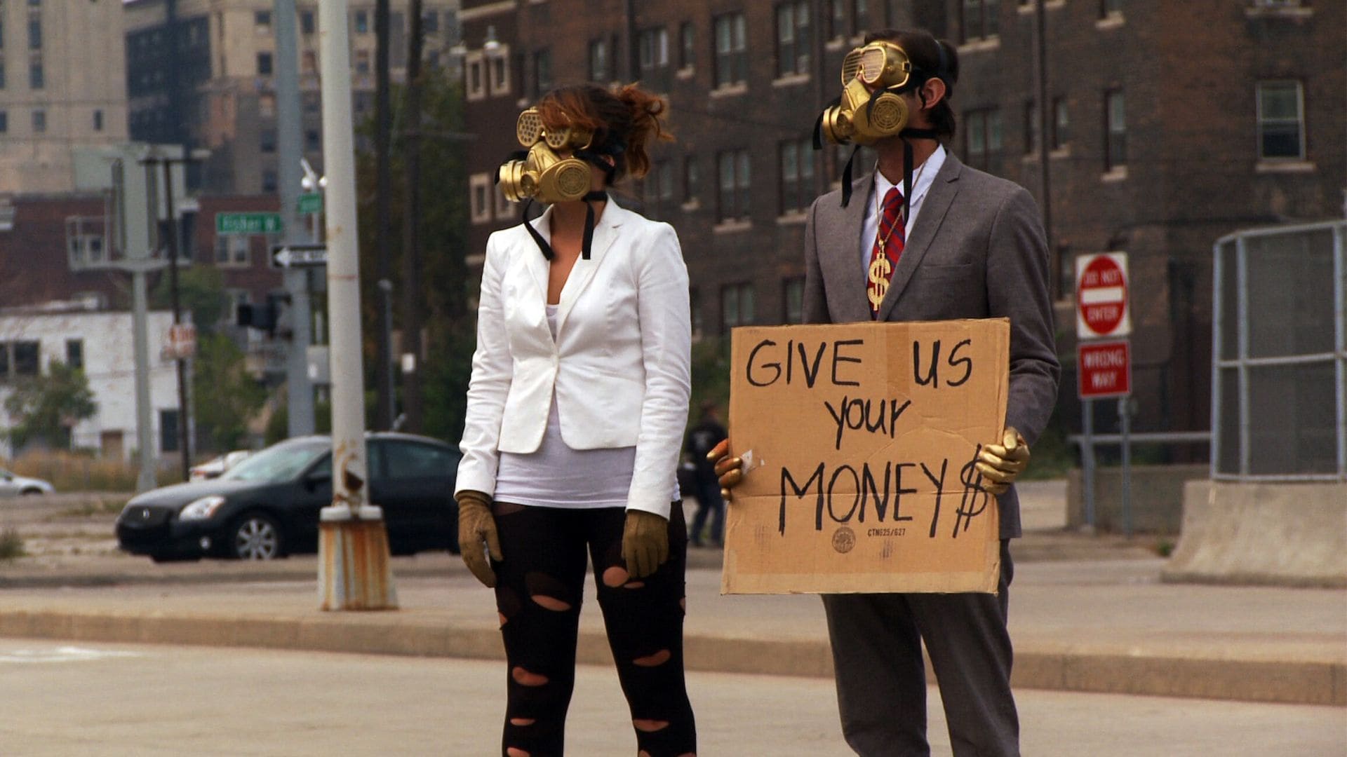 A man and woman dressed in gold plated radiation masks hold a sign that says: Give us your Money $.