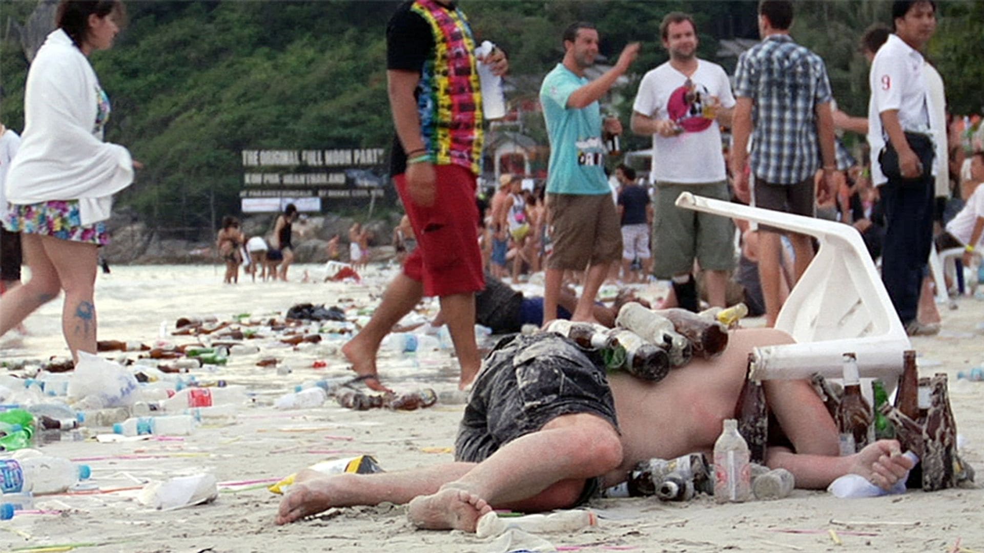 A man lays facedown in the sand at a beach; bottles of beer are scattered around and on top of him while a group of people walk around.