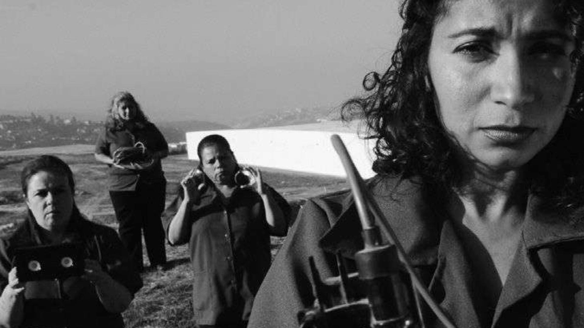 A black-and-white photo of four women in jumpsuits, with a city in the distance.