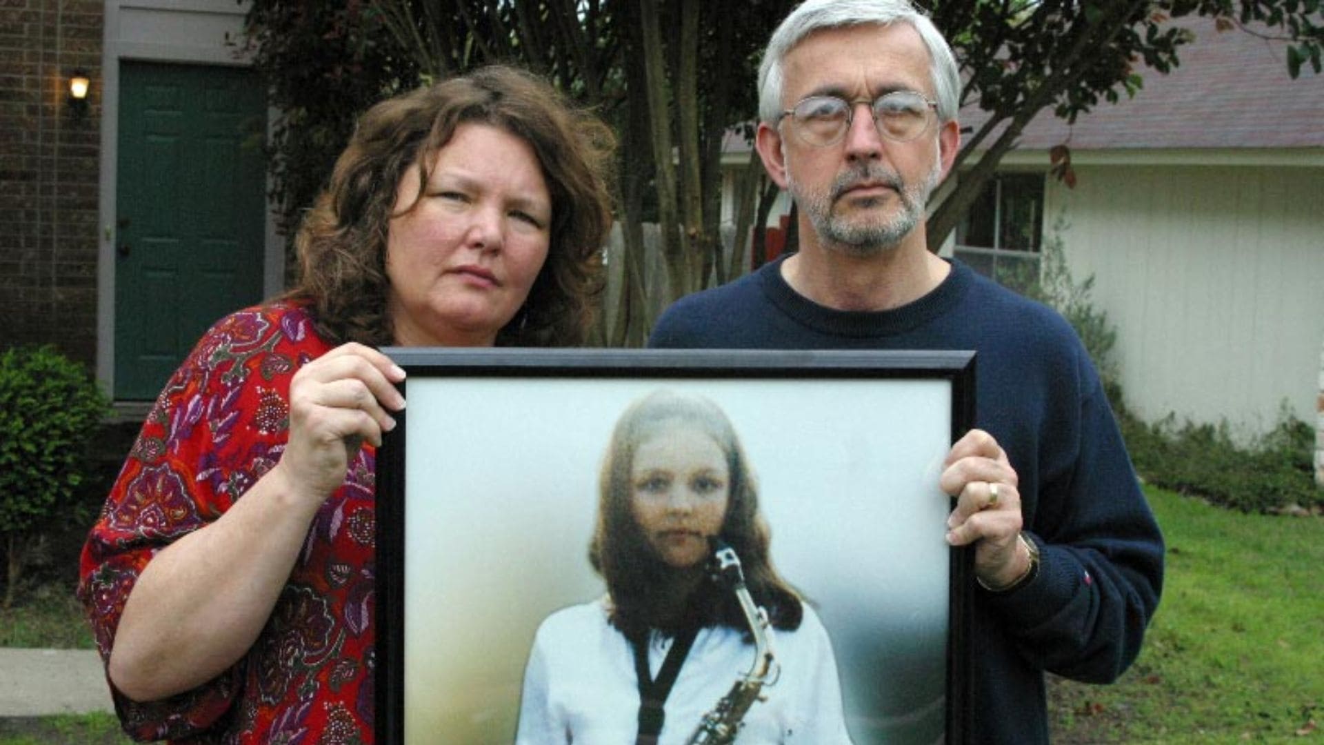 A man and a woman hold a framed picture of their daughter with an instrument.