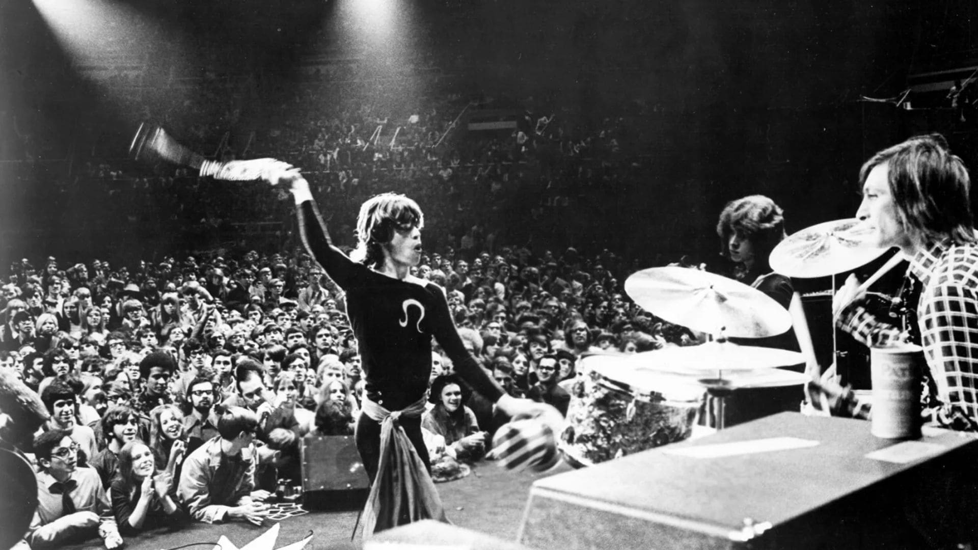 Musician Mick Jagger swings a microphone onstage in front of his band and a crowd of people.